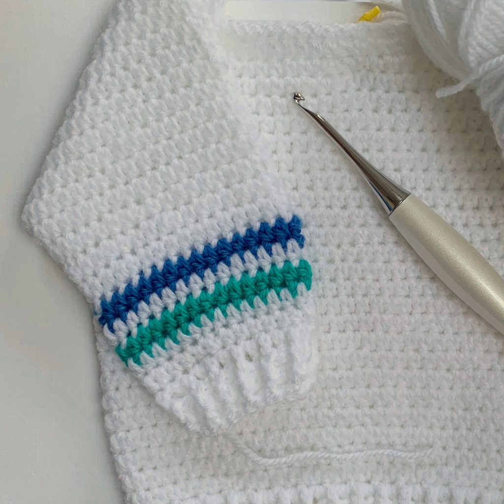 white crochet baby sweater with stripes on sleeves; crochet hook lying on top